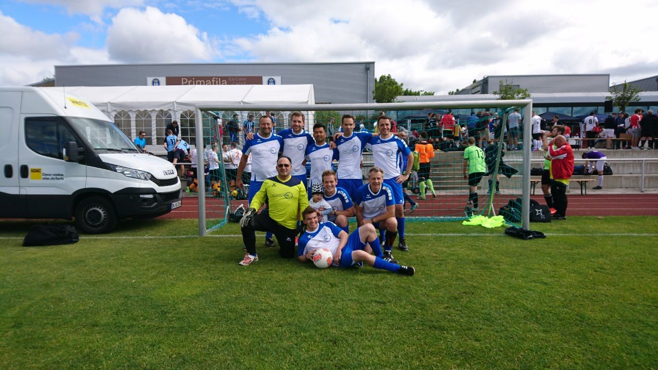 Eine Firmenfußballmannschaft in blau-weißen Uniformen posiert gemeinsam vor einem Tor auf einem Rasenplatz, im Hintergrund sind ein Lieferwagen und Zuschauer zu sehen.