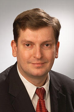 A man in a suit and red tie poses for a professional headshot against a light grey background, ready to lead successful warehouse collaboration initiatives.
