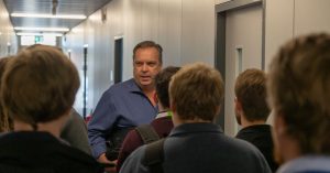 Ein Mann in einem blauen Hemd steht in einem Flur und spricht mit einer Gruppe von Menschen, die der Kamera den Rücken zuwenden.