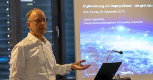 Ein Mann im weißen Hemd steht neben einer projizierten Präsentationsfolie über die Digitalisierung der Lieferkette beim SAE Institute, spricht und gestikuliert mit einer Hand.