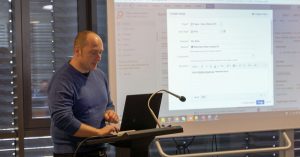 Ein Mann steht mit einem Laptop an einem Podium, hinter ihm wird auf einem projizierten Computerbildschirm ein Formular zur Problemverfolgung angezeigt.