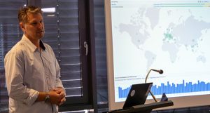 Ein Mann steht neben einem Bildschirm, auf dem eine Weltkarte mit Datenpunkten und einem Balkendiagramm angezeigt wird, und präsentiert einem Publikum im SAE Institute Informationen.