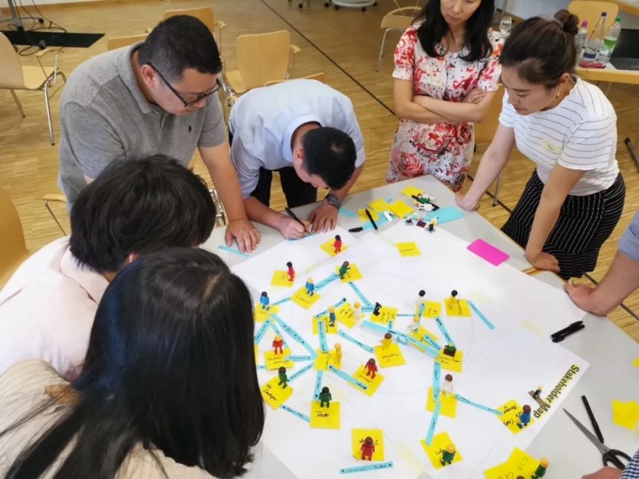 Seven people gather around a table covered with diagrams, sticky notes, and small figurines, collaborating and discussing in a brightly lit room.
