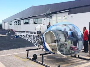 A small helicopter with a bubble-shaped cockpit is parked on the tarmac near a large hangar, with several people standing nearby.