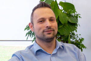 A man with short dark hair and a trimmed beard, wearing a light blue shirt, sits in front of green leafy plants and looks at the camera, appearing ready to discuss Product Owner tasks.