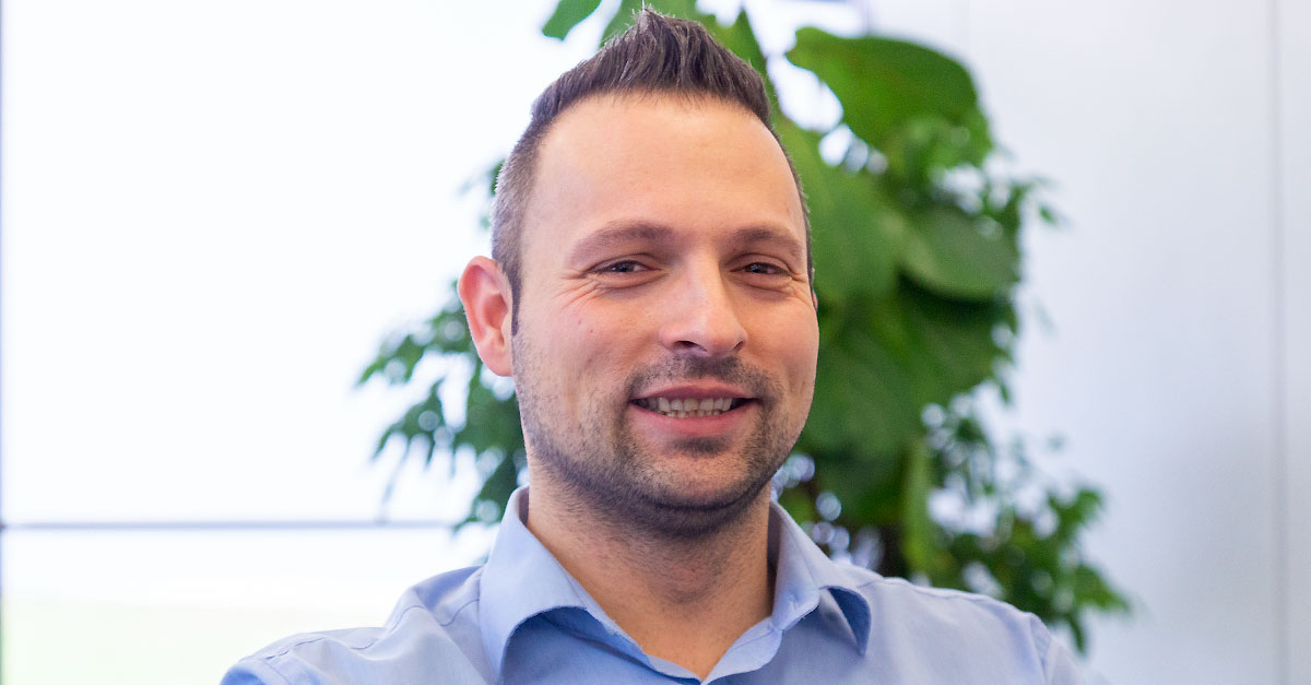 A man with short dark hair and a trimmed beard, wearing a light blue collared shirt, smiles in an indoor setting with a leafy plant in the background, suggesting confidence in handling Product Owner Tasks.