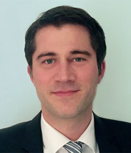 A man with short dark hair wearing a dark suit, white shirt, and striped tie stands confidently in front of a plain light background, presenting a professional image on the Materialebene.