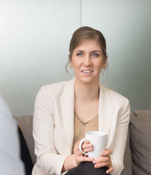 A working student in a light blazer sits on a couch holding a white mug, looking toward someone out of frame.