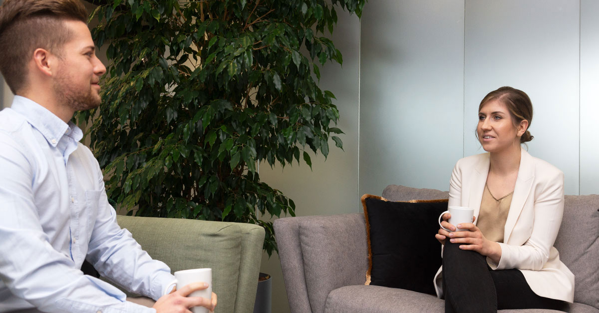 Two people, including a working student, sit on couches in an office lounge area, holding white mugs and having a conversation. A large green plant is in the background.