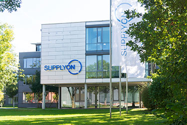 Modern office building with large glass windows and the word SUPPLYON displayed on both the facade and a flag, surrounded by trees and greenery.