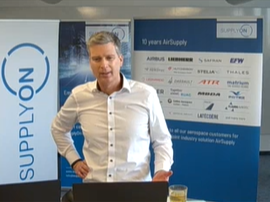 A man stands in front of SUPPLYON banners displaying various company logos, giving a presentation at Supplier Community Day. A laptop and a glass are on the table in front of him.