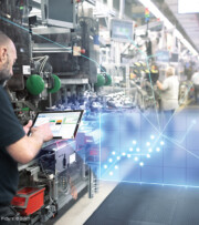 A man uses a tablet on a factory floor with machinery in the background; a digital graph overlay indicates data analysis or monitoring.