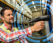 A man in a warehouse uses a handheld barcode scanner, wearing a safety vest and standing next to shelves filled with boxes. Blue circular graphics overlay the image.
