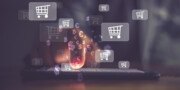 A hand interacts with a smartphone, with shopping cart and currency symbols digitally overlaid, representing online shopping and e-commerce.