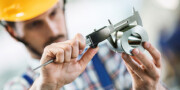 A person wearing a yellow hard hat measures a metal part with a digital caliper, focusing closely on the measurement.