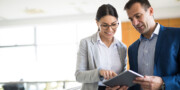 Two business professionals, a woman and a man, review information together on a digital tablet in a modern office setting.