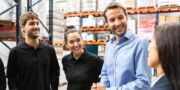 Four people stand and smile while talking together in a warehouse setting, with shelves and boxes visible in the background.