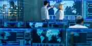 People in a control room monitor global digital maps and data on large screens, with multiple computer stations and monitors displaying similar information.