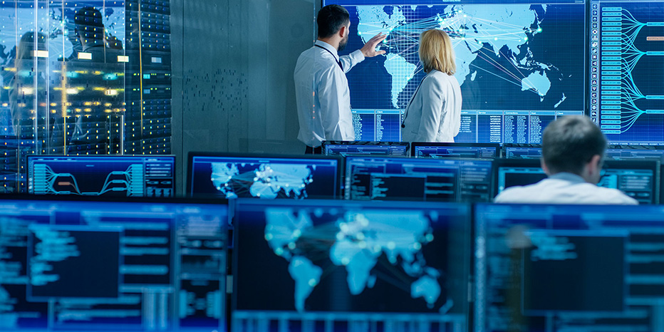 People in a control room monitor global digital maps and data on large screens, with multiple computer stations and monitors displaying similar information.