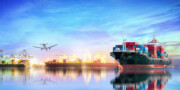 Container ships dock at a busy port with cranes unloading cargo; an airplane flies overhead against a colorful sky reflected in calm water.