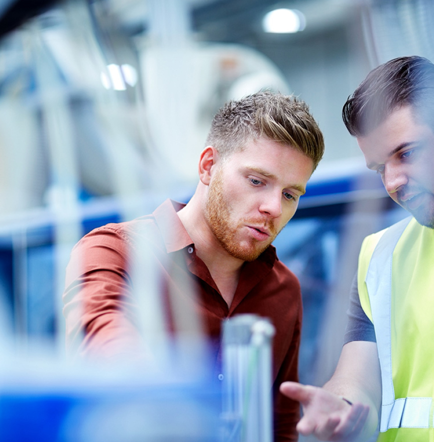 Two men discuss machinery in an industrial setting; one wears a brown shirt, the other a yellow safety vest.