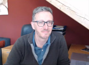 A man with glasses sits at a desk in an office with a red wall and wooden ceiling, facing a computer screen.