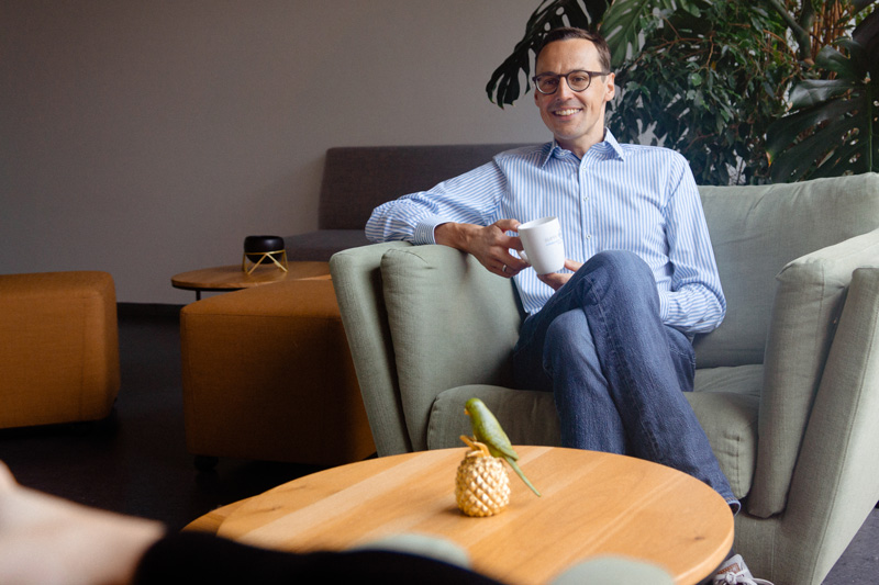 A man in glasses sits on a light green armchair, enjoying his Teilzeit break, holding a white mug with plants and a table featuring a pineapple decoration in front of him.