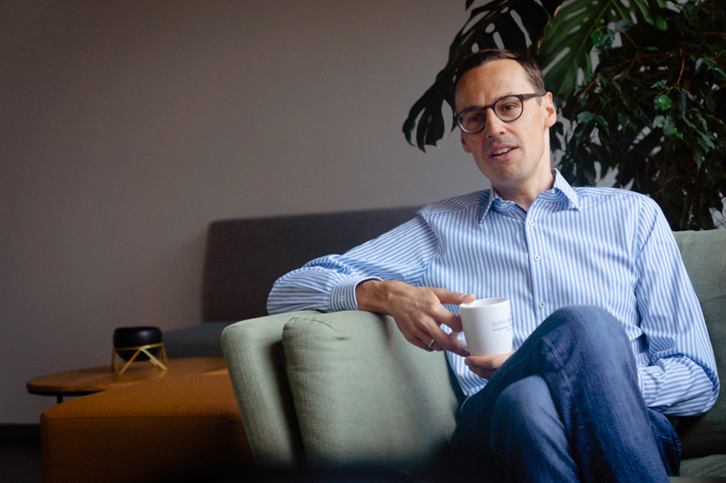 A part-time worker in glasses and a striped shirt sits on a green sofa, holding a white mug, with indoor plants and modern furniture in the background.
