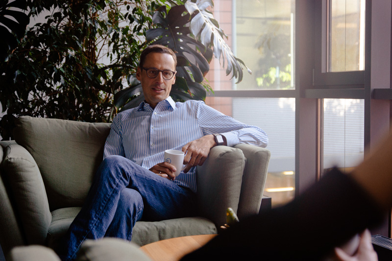 A part-time worker wearing glasses and a striped shirt sits on a couch holding a cup, with crossed legs, in a well-lit room filled with large windows and plants.