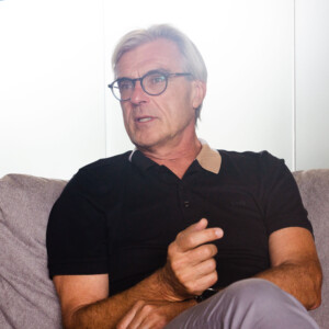 An older man with gray hair and glasses sits on a couch, wearing a black polo shirt and gesturing with his hand as he discusses the importance of connectivity.