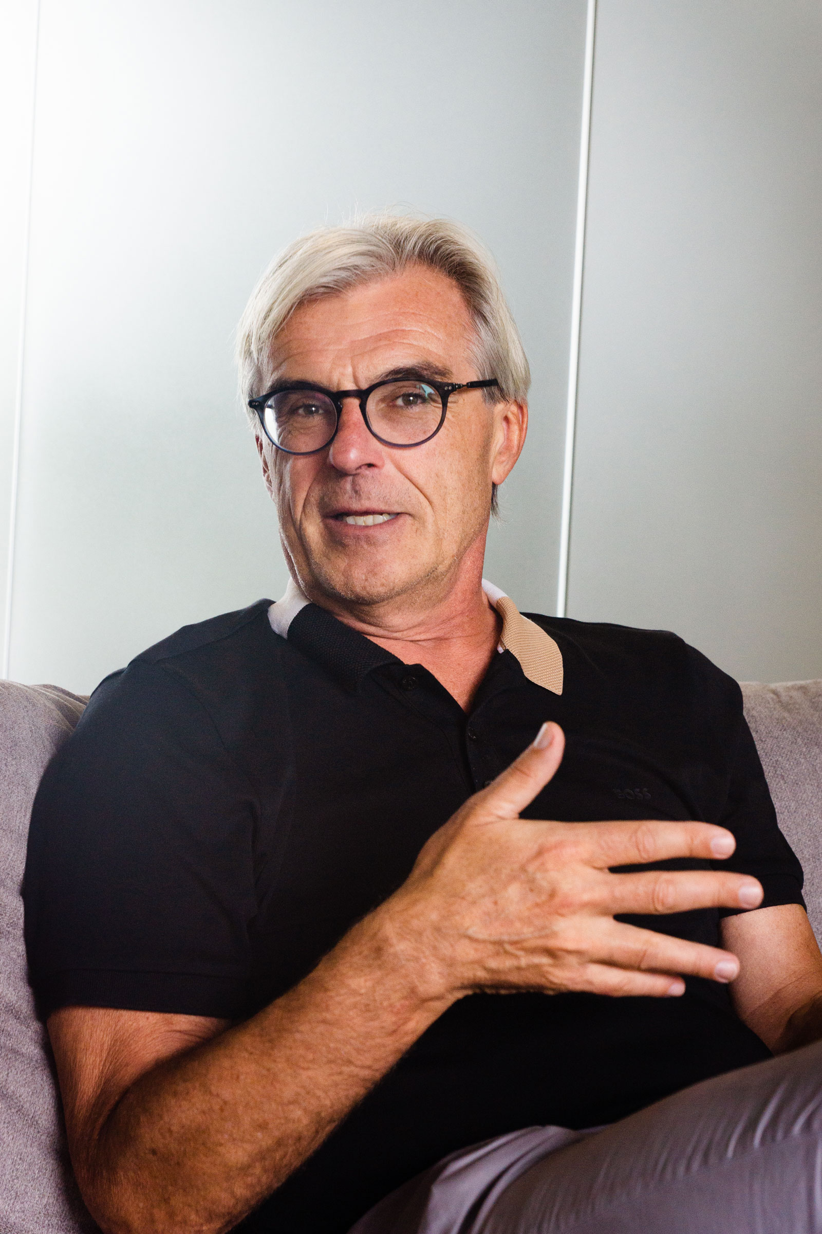 A middle-aged man with gray hair and glasses sits on a couch, wearing a black polo shirt, and gestures with his right hand while talking about the importance of connectivity.