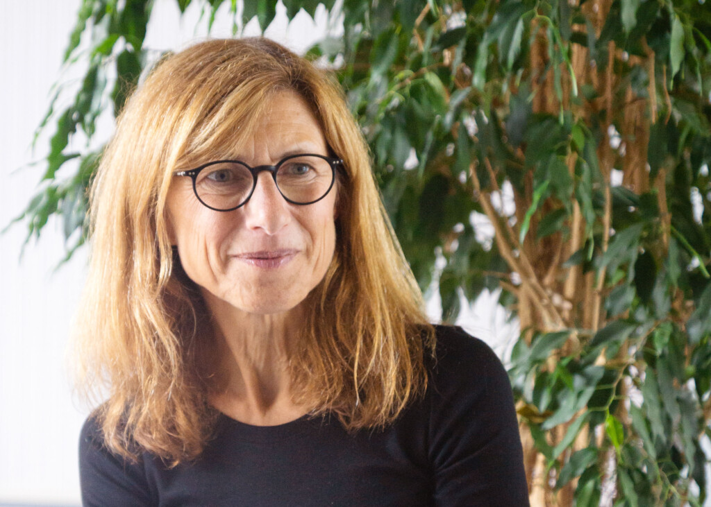A woman with shoulder-length light brown hair and glasses, wearing a black top, sits in front of a leafy green plant, embodying a sense of calm and connectivity with nature.