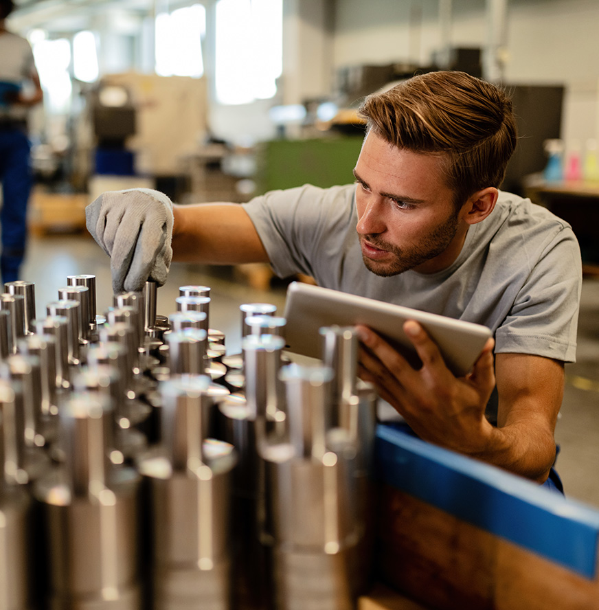 A man wearing a glove inspects metal cylindrical parts while holding a tablet, utilizing digital twin technology to simplify traceability and improve product quality in an industrial setting.