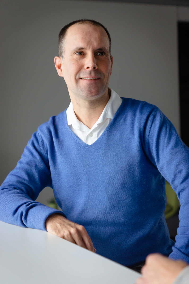 A man in a blue sweater and white collared shirt sits at a table, smiling and looking slightly to the side—perhaps discussing the latest updates to the Supply Chain Act.