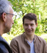 Two people sit outdoors having a conversation; one is an older man with glasses, and the other is a young man smiling. Greenery is visible in the background.