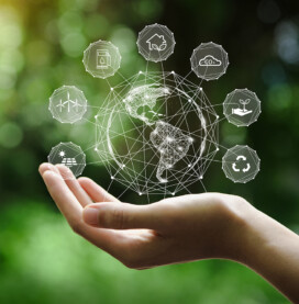 A hand holds a digital globe with interconnected eco-friendly icons, representing concepts like recycling, renewable energy, and sustainable development against a blurred natural background.