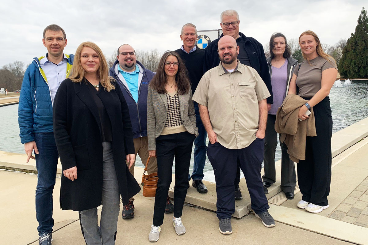 A group of nine adults, attending a conference, stand together outdoors near a reflecting pool on a cloudy day, posing for a group photo.