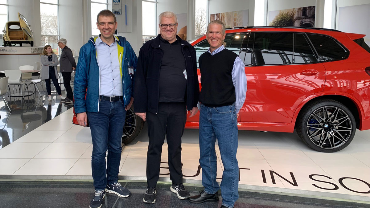 Three men stand in front of a red SUV inside a modern showroom, possibly attending a conference, with large windows and other people in the background.