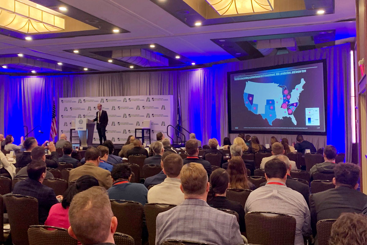 A speaker presents to an audience at a conference in a room, showing a map of the United States with highlighted states on a large screen.