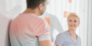 A man and a woman stand by a whiteboard with sticky notes, engaged in conversation. The woman is smiling and looking at the man.