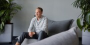 A man sits on a gray sofa in a modern room with potted plants, wearing a light shirt and dark pants, looking slightly to the side.