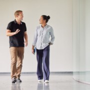 A man and a woman walk side by side in a modern hallway, engaged in conversation, with both smiling and dressed in casual business attire.