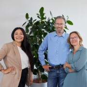 Three adults stand together in front of a large indoor plant, smiling at the camera in a brightly lit room.