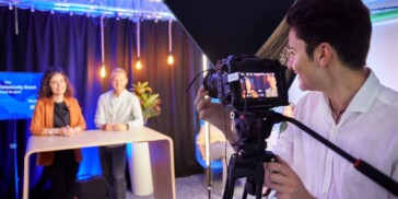 A person operates a camera recording two people standing behind a table in a brightly lit studio with a screen and decorative lights.