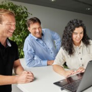 Three people are gathered around a laptop, looking at the screen and smiling in a modern office setting with a plant in the background.