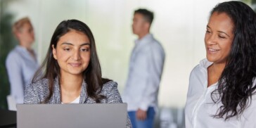 Two women are looking at a laptop together in the foreground, while two people stand and talk in the blurred background of an office setting.