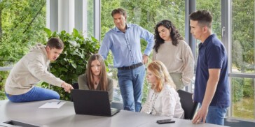 Six people gather around a table in an office, looking at a laptop screen. One person is pointing at the laptop while others observe attentively. Large windows show greenery outside.