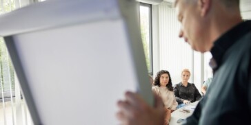 A man stands by a blank flip chart, speaking to three people seated at a table in a well-lit meeting room.
