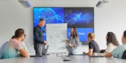 A group of people in a meeting room watch two presenters explaining a diagram on a flipchart, with digital twin graphics displayed on screens behind them.