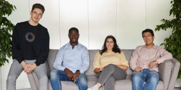 Four people, three seated on a gray couch and one standing next to it, pose for a group photo in a bright room with green plants.